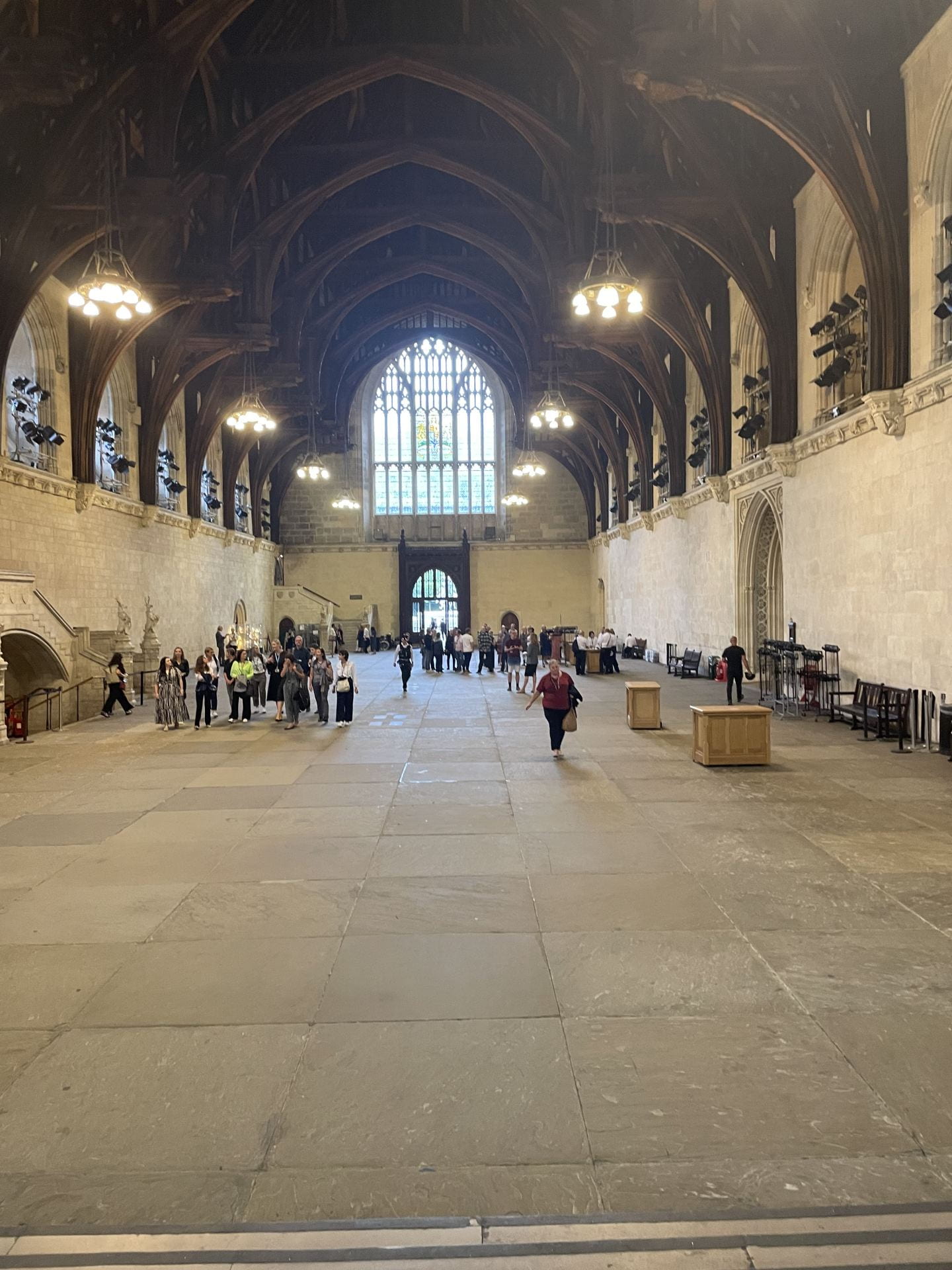An image of Westminster Hall taken from one end to show the full length of the grand building. It has dark wooden beamed structures in the roof space and the floor is grey composed of many square slate slabs. There are many visitors in the space
